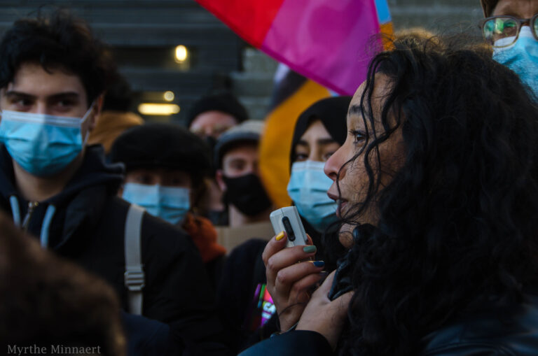Stevie spreekt in de microfoon van een megafoon omringd door verschillende mensen met mondkapjes op een demonstratie.